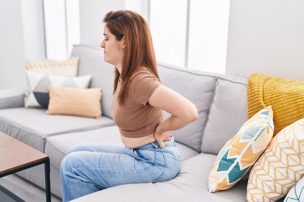 Woman sitting on a couch holding her lower back due to pain and discomfort at home